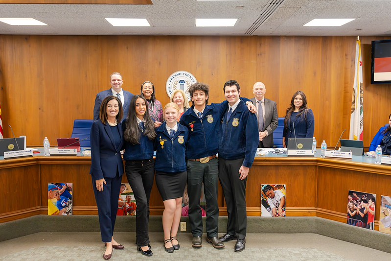 A group of students pose with Fresno County Superintendent of School and Board of Education education leaders during a board meeting, standing in front of the board dais.