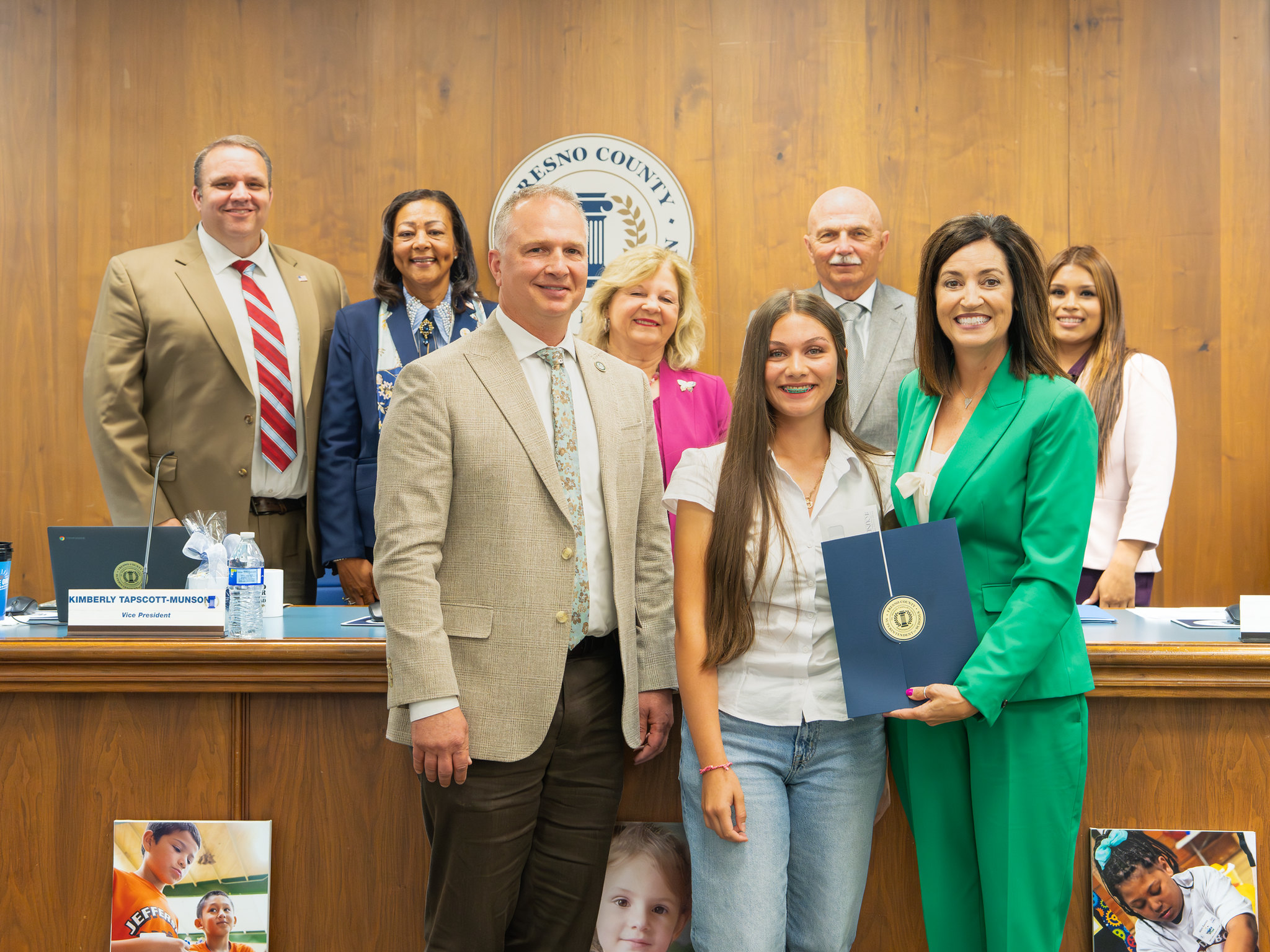 A group of nine adults and one student stand together in a formal boardroom setting, smiling for a photo. At the front, a teenage girl holds a certificate while standing between the Superintendent of Schools, Dr. Michele Copher in green and the District Superintent, Jeff Purcell. Behind them is the Board of Education in front of a wooden wall featuring a county seal.