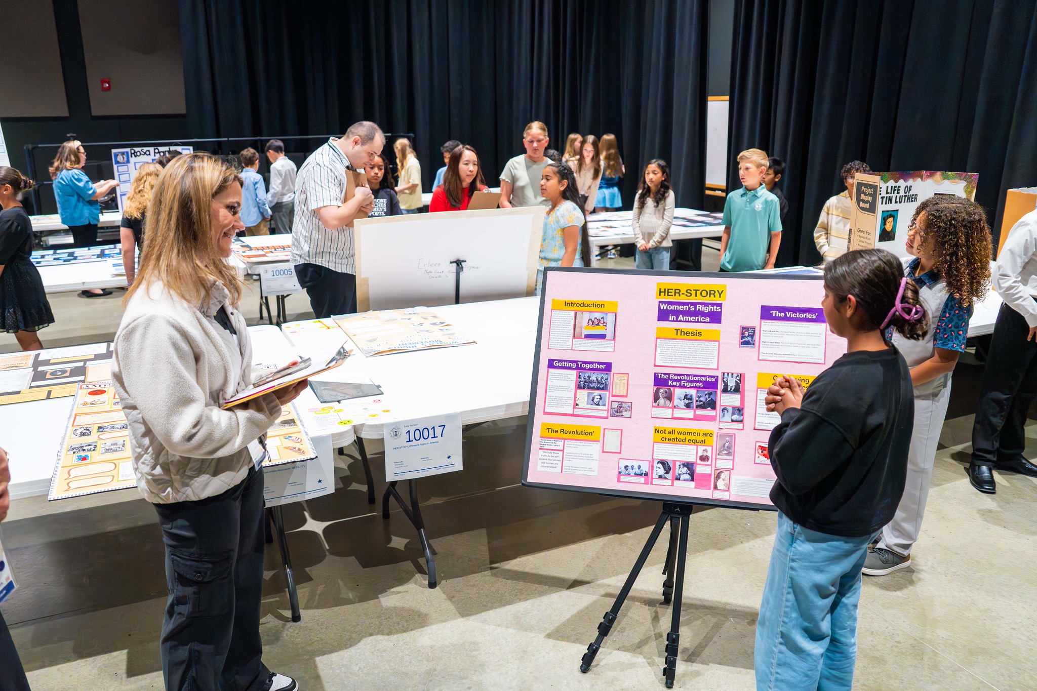 Student stands beside her project display while a judge evaluates it, surrounded by other students presenting their work in a busy room.