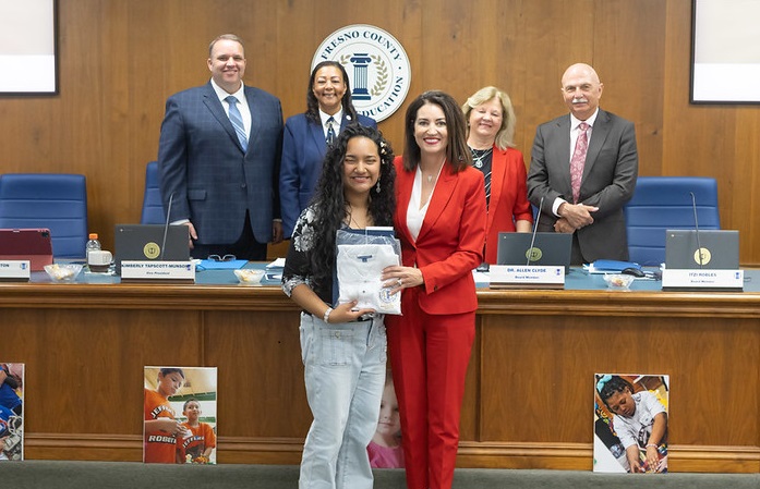 A student poses with the Superintendent, Dr. Michele Copher and the Board of Education standing behing them during a board meeting.